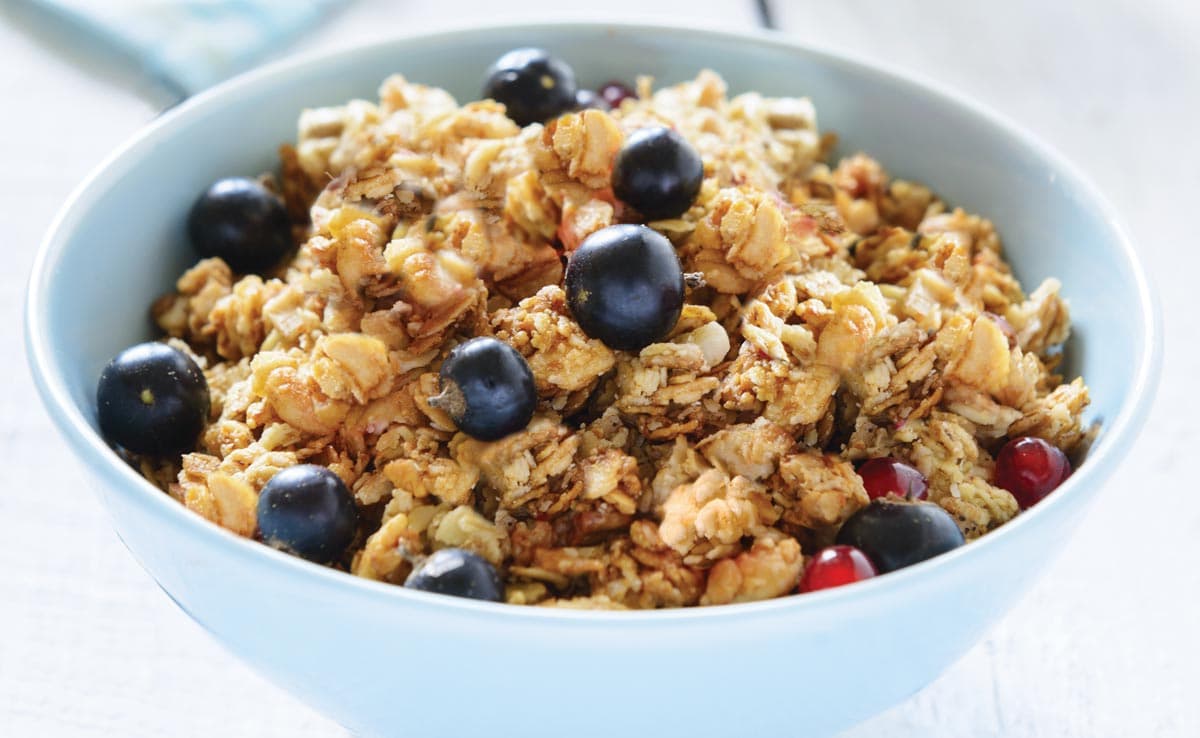 A light blue bowl filled with granola and topped with fresh blueberries and red berries sits on a white surface.