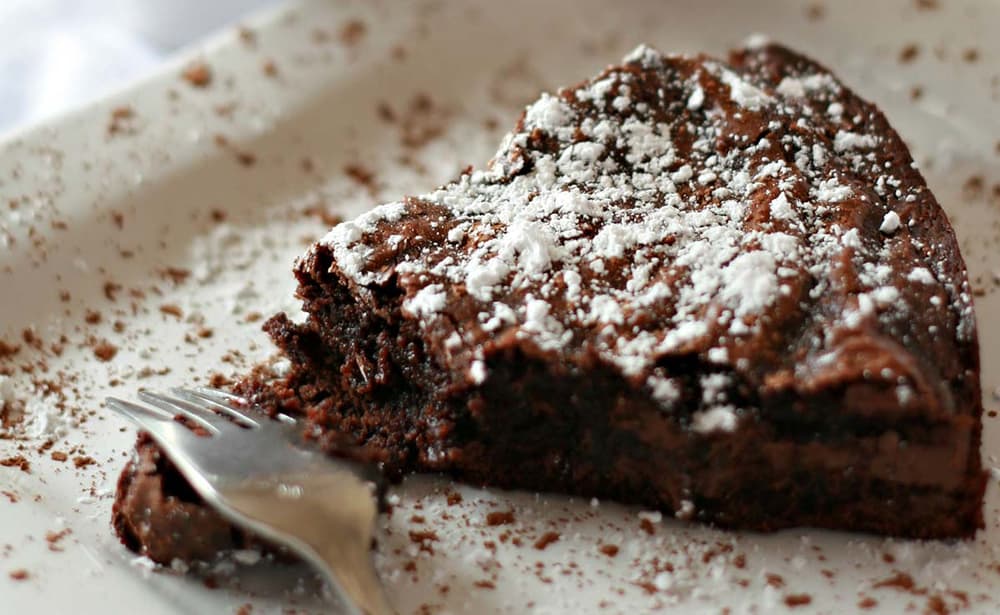 A partially eaten slice of chocolate cake topped with powdered sugar sits on a white plate next to a fork.