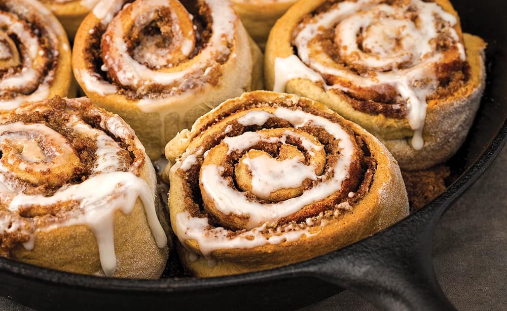 Cinnamon rolls with icing are arranged closely together in a cast iron skillet, showing swirled layers and melted glaze.