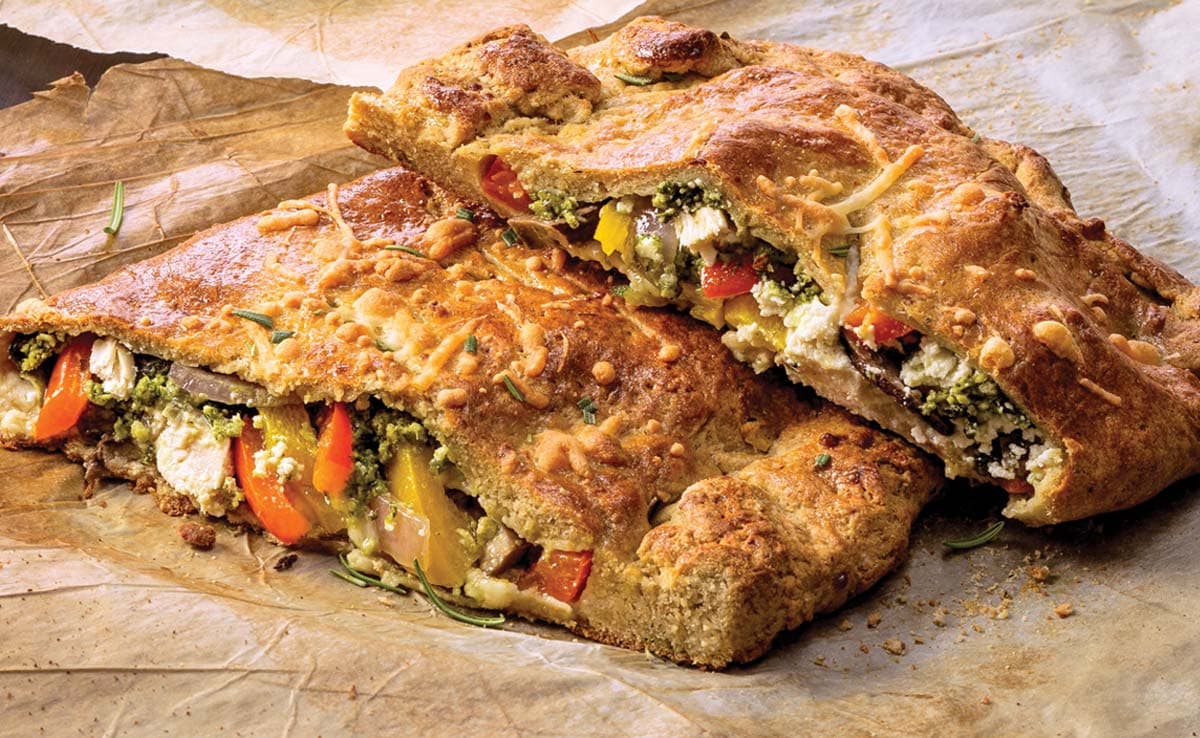 Two slices of vegetable-stuffed calzone with a golden-brown crust, showing colorful bell peppers, cheese, and green pesto on parchment paper.