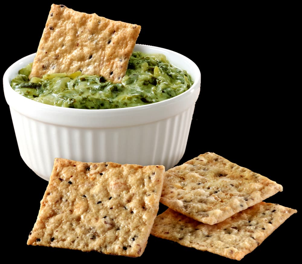 A white ramekin filled with green dip, with a cracker inside, is placed next to three square crackers on a white surface.