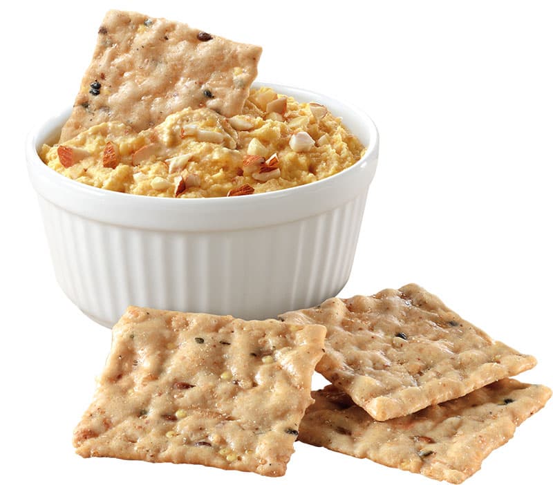 A white ramekin filled with hummus, topped with chopped nuts, with several multigrain crackers beside and one cracker dipped in the hummus.