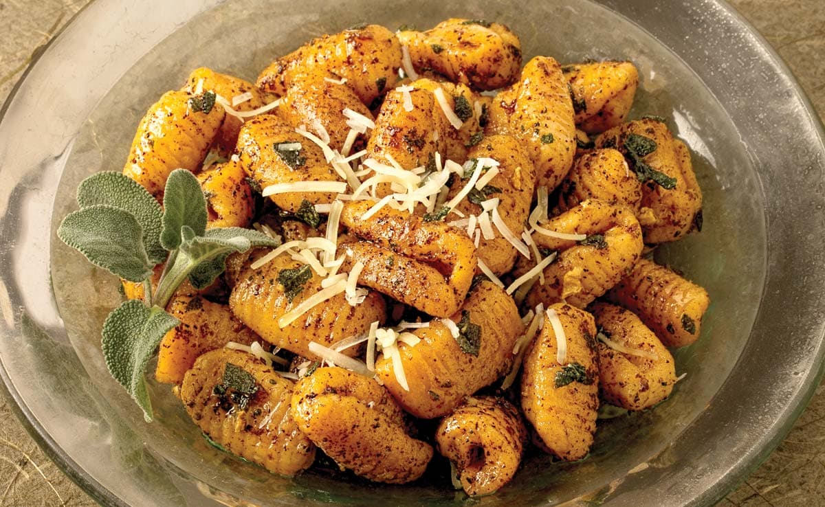 A glass bowl filled with pumpkin gnocchi topped with grated cheese, browned butter, herbs, and garnished with fresh sage leaves.