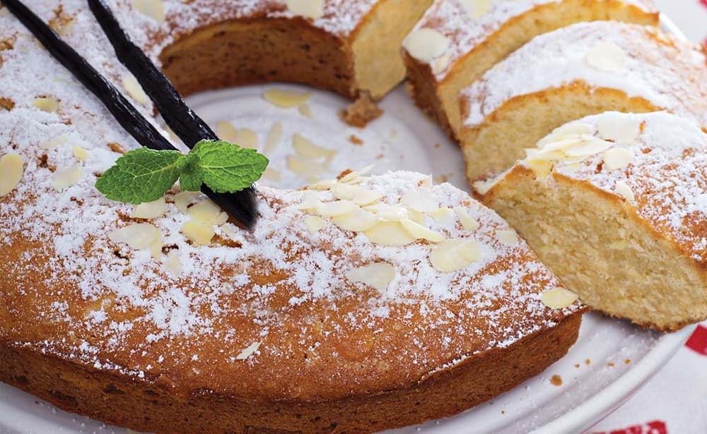 Round almond cake dusted with powdered sugar, garnished with sliced almonds, mint leaves, and two vanilla pods on a white plate. One slice is removed.