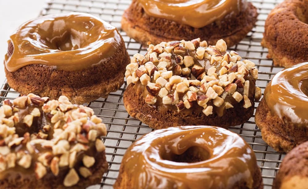 Baked donuts topped with caramel glaze and chopped nuts are cooling on a wire rack.