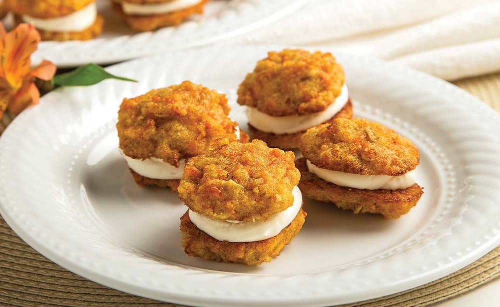 Four carrot cake whoopie pies with cream cheese filling are arranged on a white plate, with more treats blurred in the background.