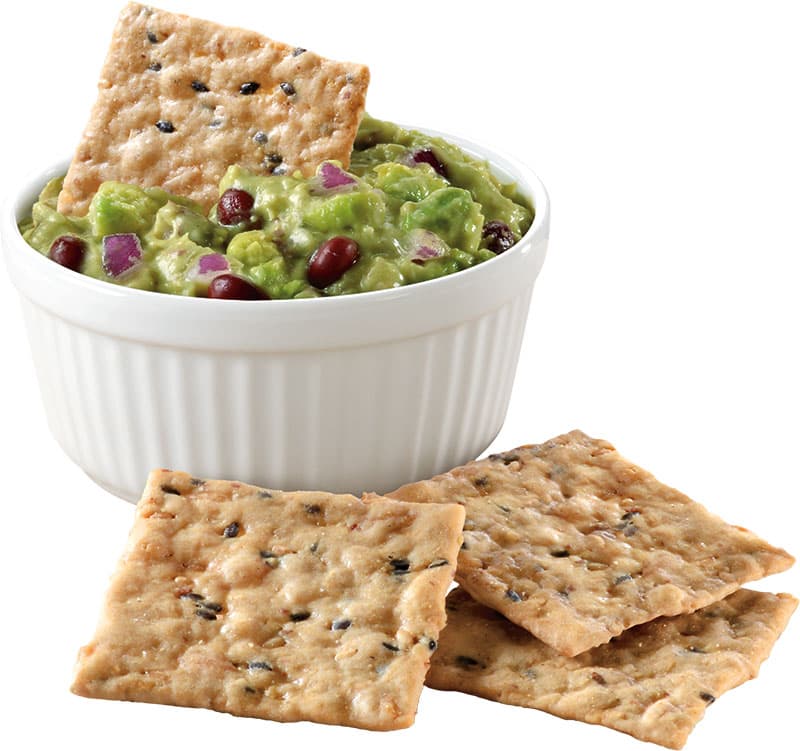 A white ramekin filled with guacamole, topped with diced red onion and beans, with several multi-grain crackers around it and one cracker dipped in the guacamole.