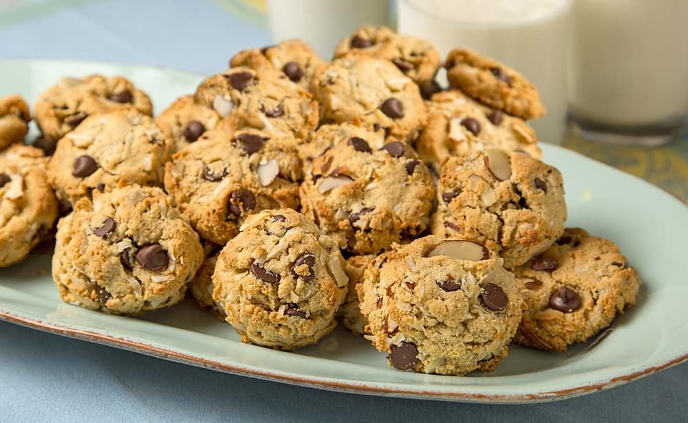 A plate of chocolate chip cookies with nuts, arranged on a light-colored platter, with two glasses of milk in the background.
