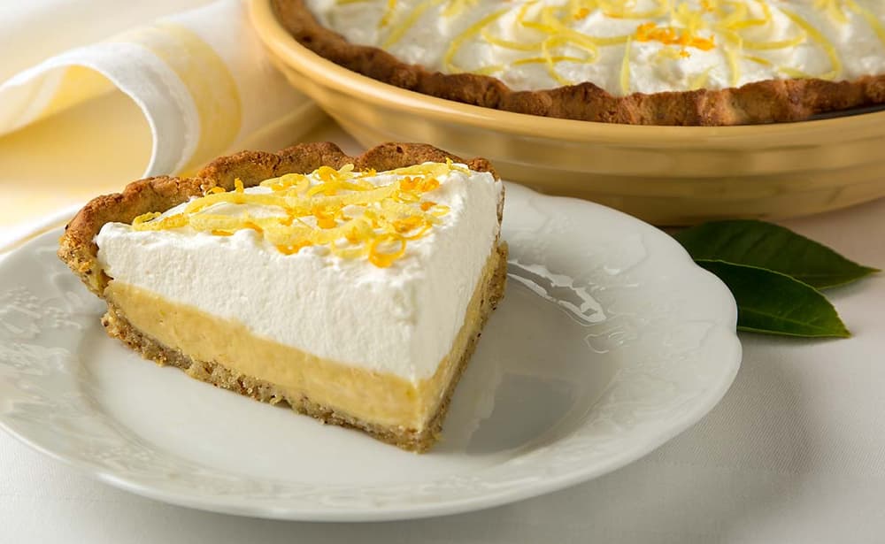 A slice of lemon cream pie with whipped topping and lemon zest sits on a white plate, with the rest of the pie in the background.