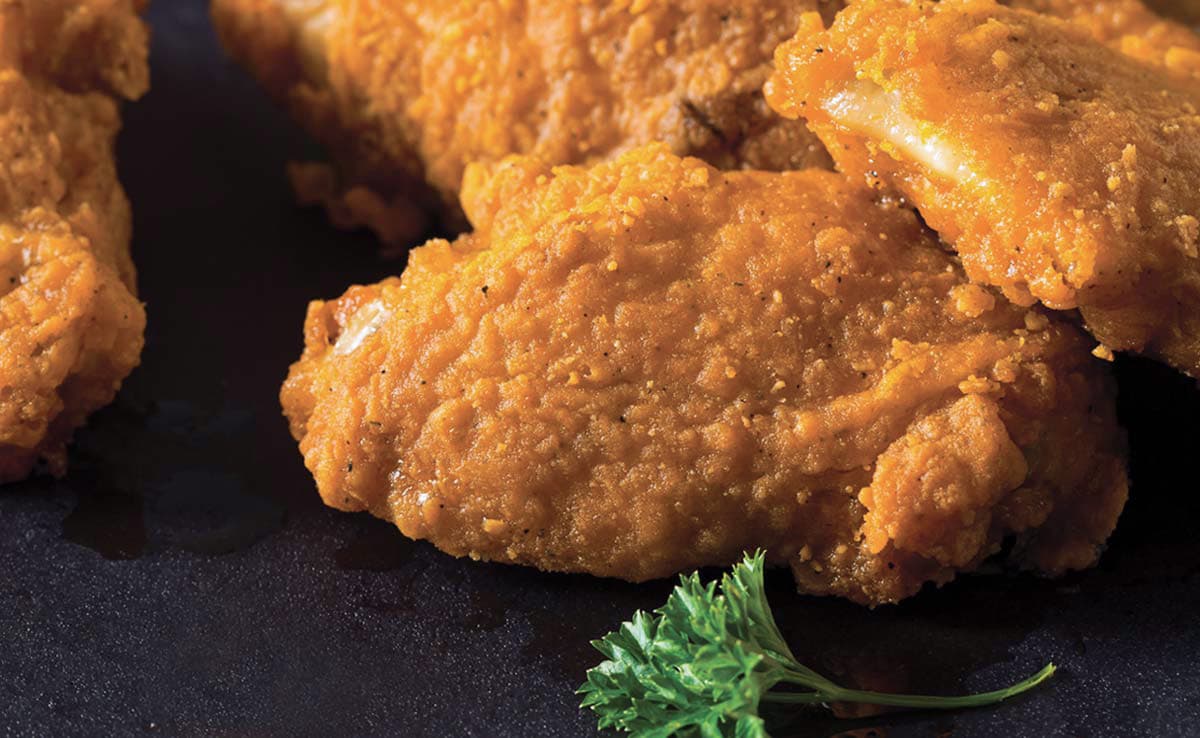 Close-up of crispy fried chicken pieces on a dark surface with a small sprig of parsley in the foreground.