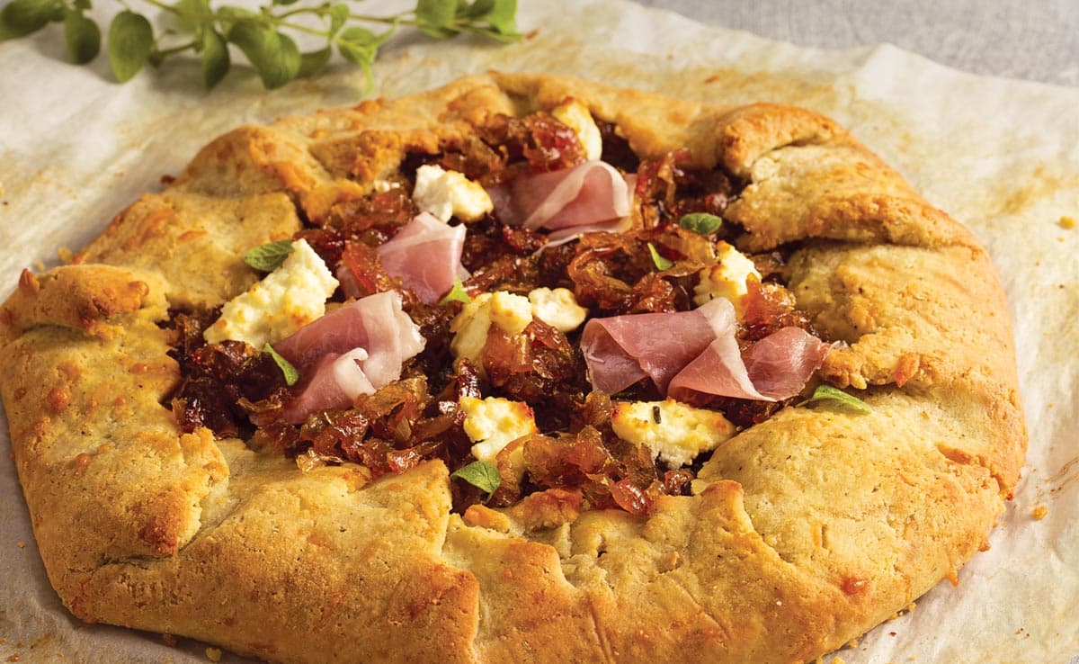 A rustic galette with a golden-brown crust, topped with caramelized onions, prosciutto, ricotta cheese, and fresh herbs, on parchment paper.