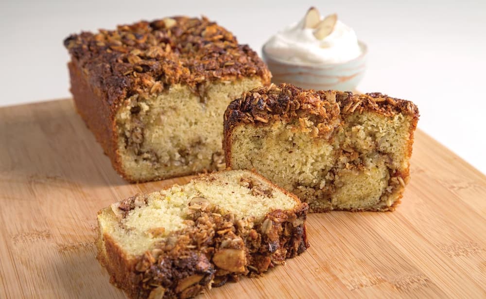 A loaf of nut bread with two slices cut, showing a nut-filled interior, on a wooden board. A small bowl of whipped cream topped with almond slices is in the background.