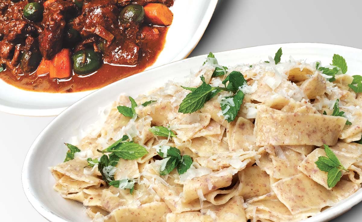 A white plate of wide, flat pasta topped with grated cheese and mint leaves sits in front of a dish of beef stew with carrots and olives.