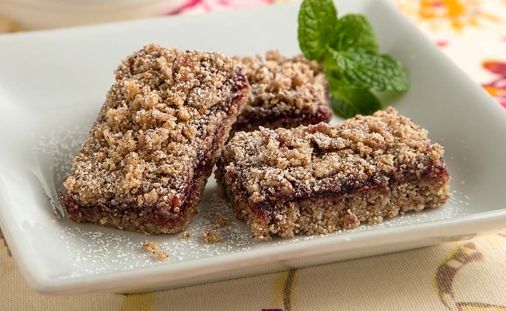 Three rectangular crumb bars with a fruit filling are arranged on a white square plate, garnished with a sprig of mint.