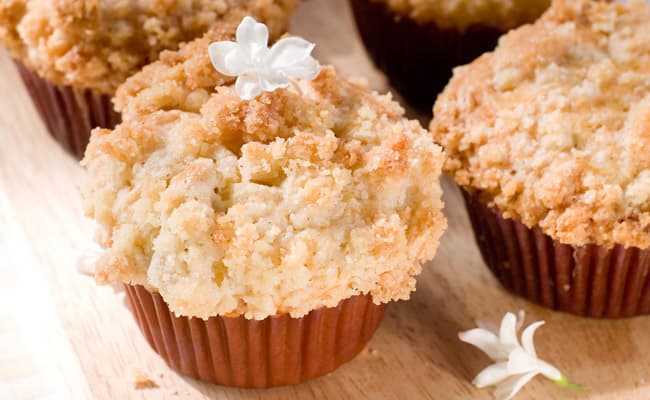 Close-up of crumb-topped muffins in brown paper liners on a wooden surface, with small white flowers as decoration.
