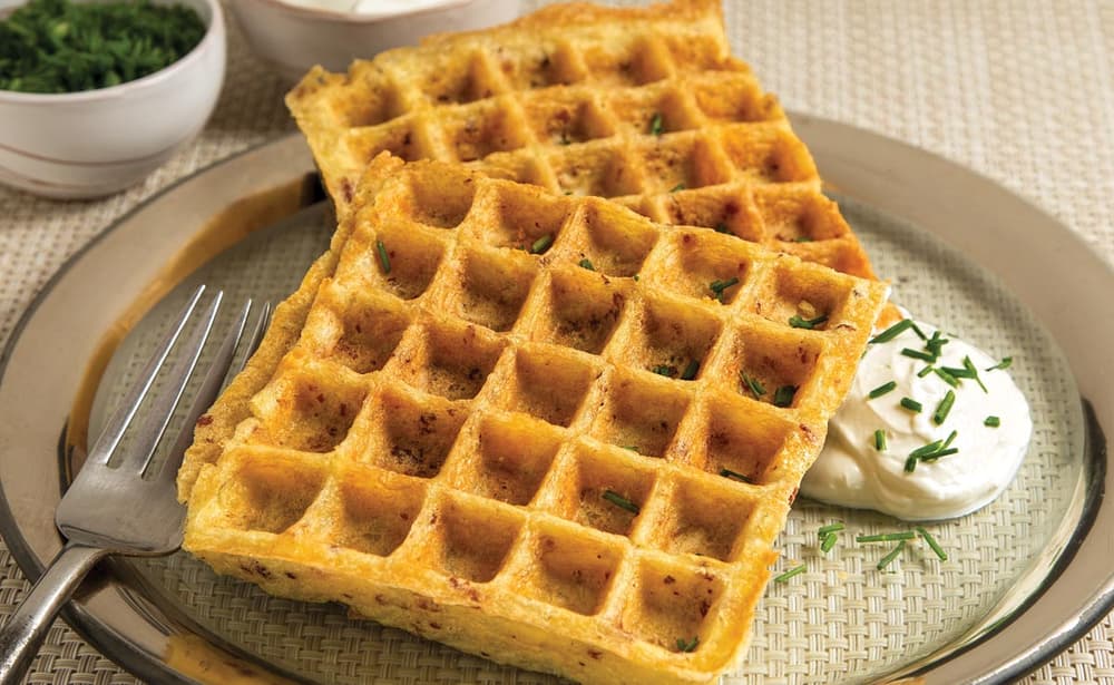 Two cooked waffles on a plate with a dollop of sour cream and chopped herbs, served with a fork on a woven placemat.