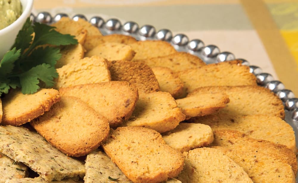 A platter of rectangular, golden-brown crackers is arranged in overlapping layers next to a small bowl of dip and a sprig of parsley.