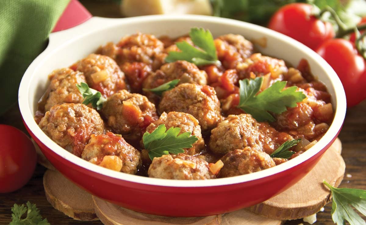 Meatballs in tomato sauce are served in a red and white pan, garnished with fresh parsley, with tomatoes and greens in the background.