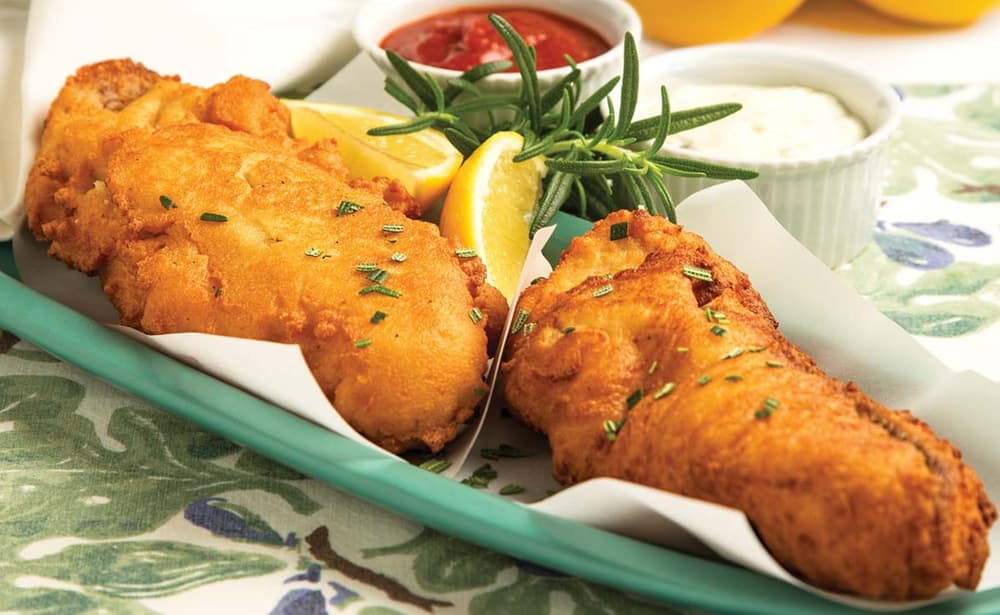 Two pieces of golden fried fish on a green plate with lemon wedges, a sprig of rosemary, and small bowls of tartar sauce and ketchup in the background.