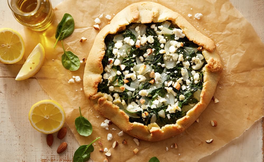 A baked spinach and cheese galette on parchment paper, surrounded by lemon wedges, spinach leaves, almonds, and a glass of olive oil.