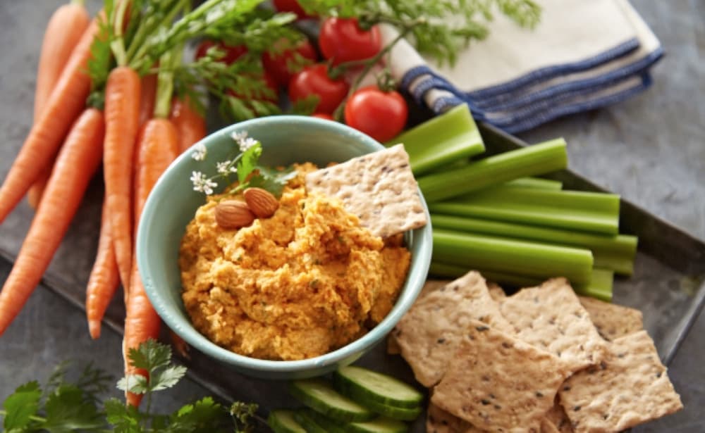 A bowl of dip with crackers, surrounded by fresh carrots, celery sticks, cherry tomatoes, cucumber slices, and herbs on a tray.