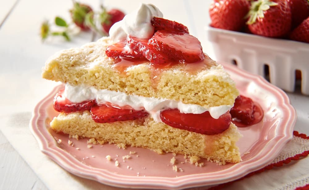 A slice of strawberry shortcake with whipped cream and strawberry slices is served on a pink plate, with fresh strawberries in the background.