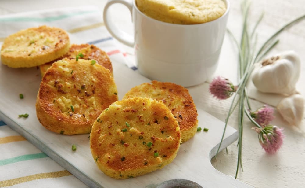 Four slices of toasted garlic bread with chopped chives on a white board, next to a mug with a baked dish, garlic bulbs, and chive flowers in the background.