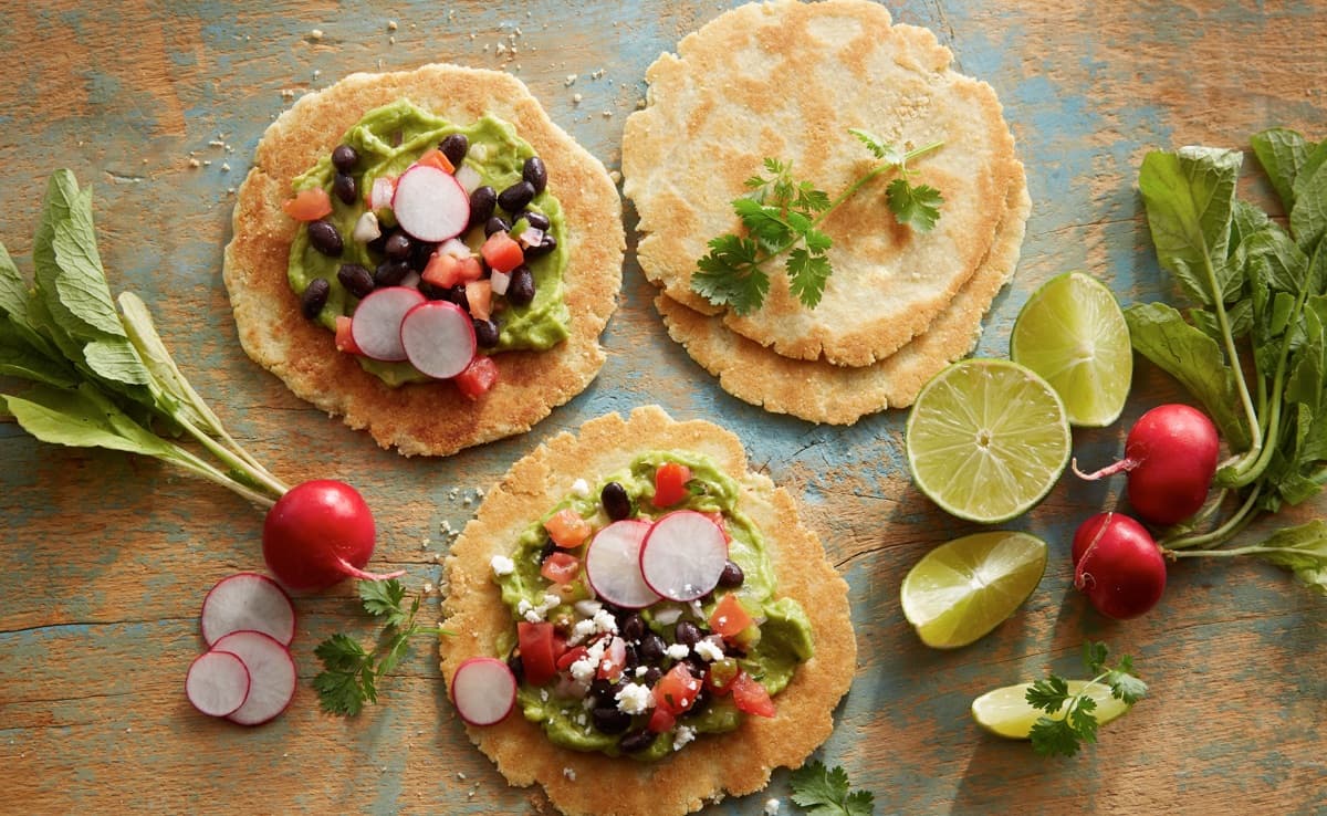 Two corn tortillas topped with guacamole, black beans, diced tomatoes, radish slices, cheese, and cilantro, with whole radishes, lime wedges, and cilantro on a rustic wooden surface.
