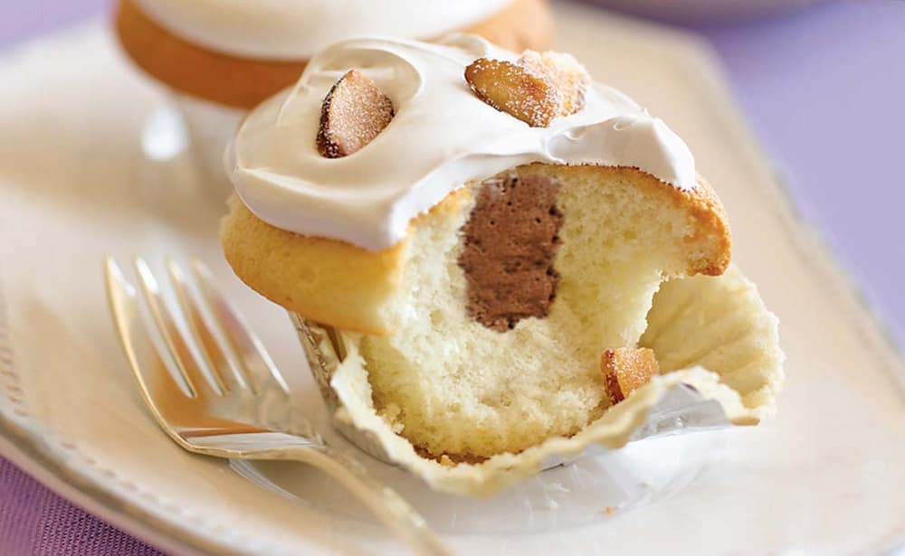 A vanilla cupcake with white icing and chocolate filling is partially eaten, sitting on a plate beside a fork.