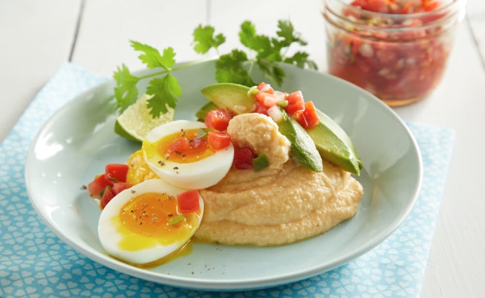 Plate with hummus, sliced avocado, soft-boiled egg, diced tomatoes, cilantro, and lime wedge; jar of salsa in the background.