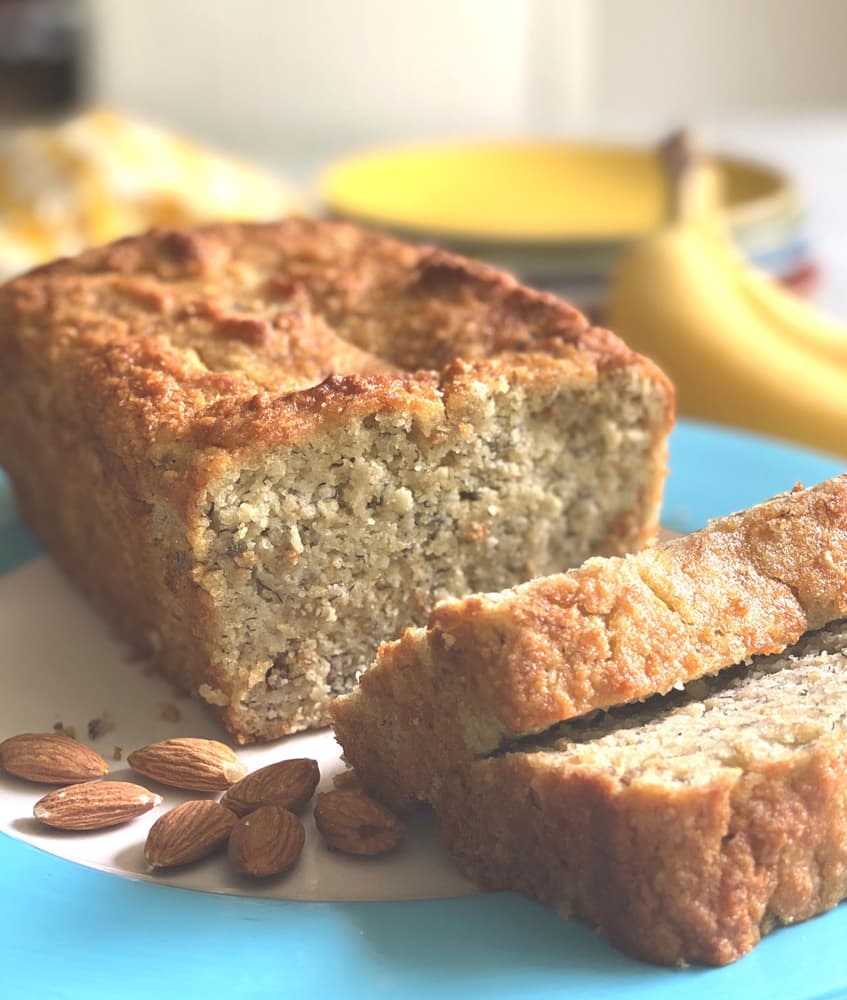 A loaf of banana bread with two slices cut, placed on a plate next to whole almonds. A banana and bowls are blurred in the background.