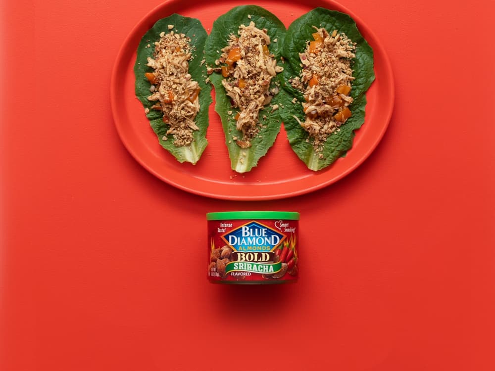 Three lettuce wraps with shredded filling on a red plate, above a can of Blue Diamond Bold Sriracha almonds, on a red background.