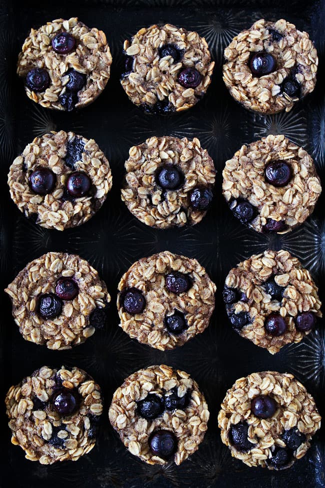 Twelve baked oatmeal cups with blueberries arranged in neat rows on a dark textured tray.