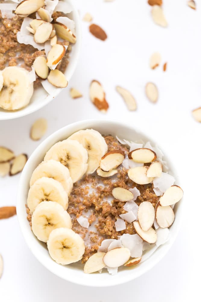 A white bowl of quinoa porridge topped with banana slices, coconut flakes, and sliced almonds. Another partial bowl and scattered toppings are visible on a white background.