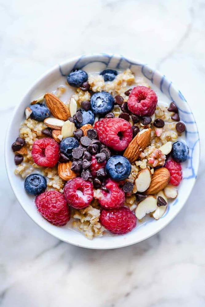 Bowl of oatmeal topped with raspberries, blueberries, sliced almonds, whole almonds, and chocolate chips, on a marble surface.