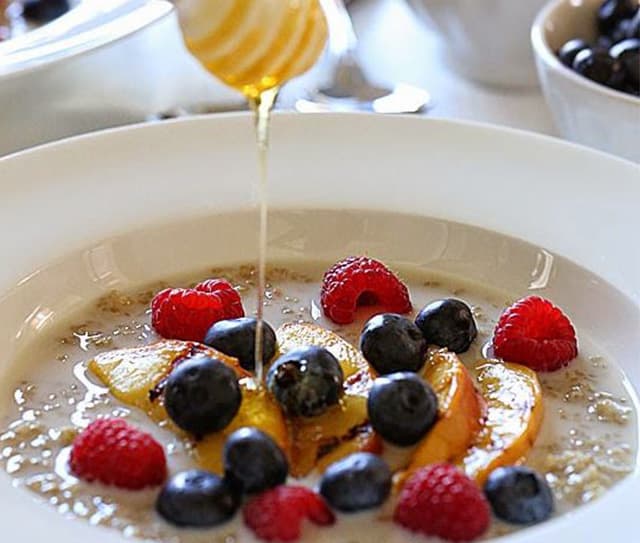 A bowl of oatmeal topped with blueberries, raspberries, and peach slices, with honey being drizzled over the fruit.