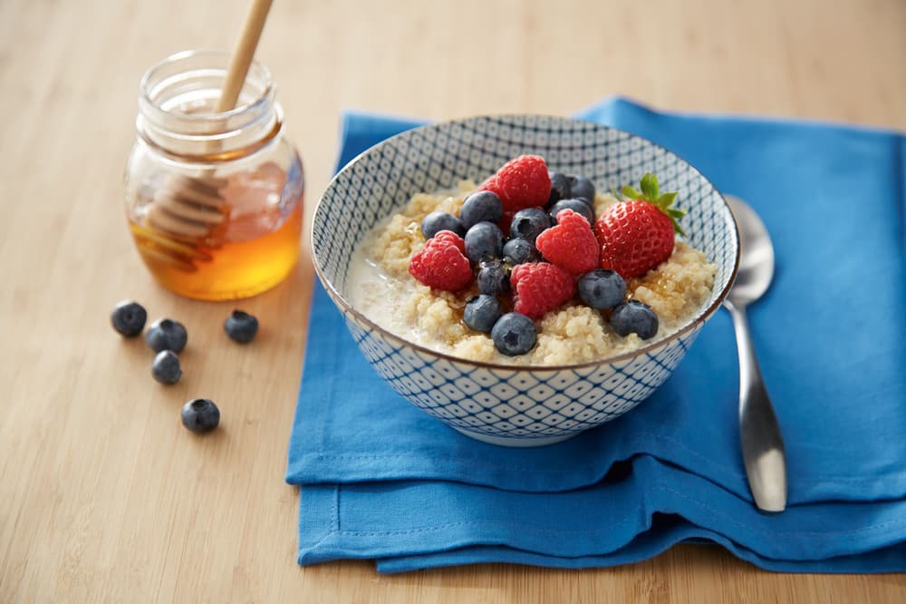 A bowl of oatmeal topped with strawberries and blueberries sits on a blue napkin next to a jar of honey, a spoon, and a few loose blueberries on a wooden surface.