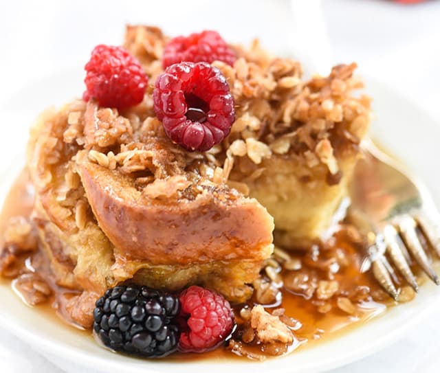 A serving of baked French toast topped with granola, raspberries, a blackberry, and drizzled with syrup, shown with a fork on a white plate.