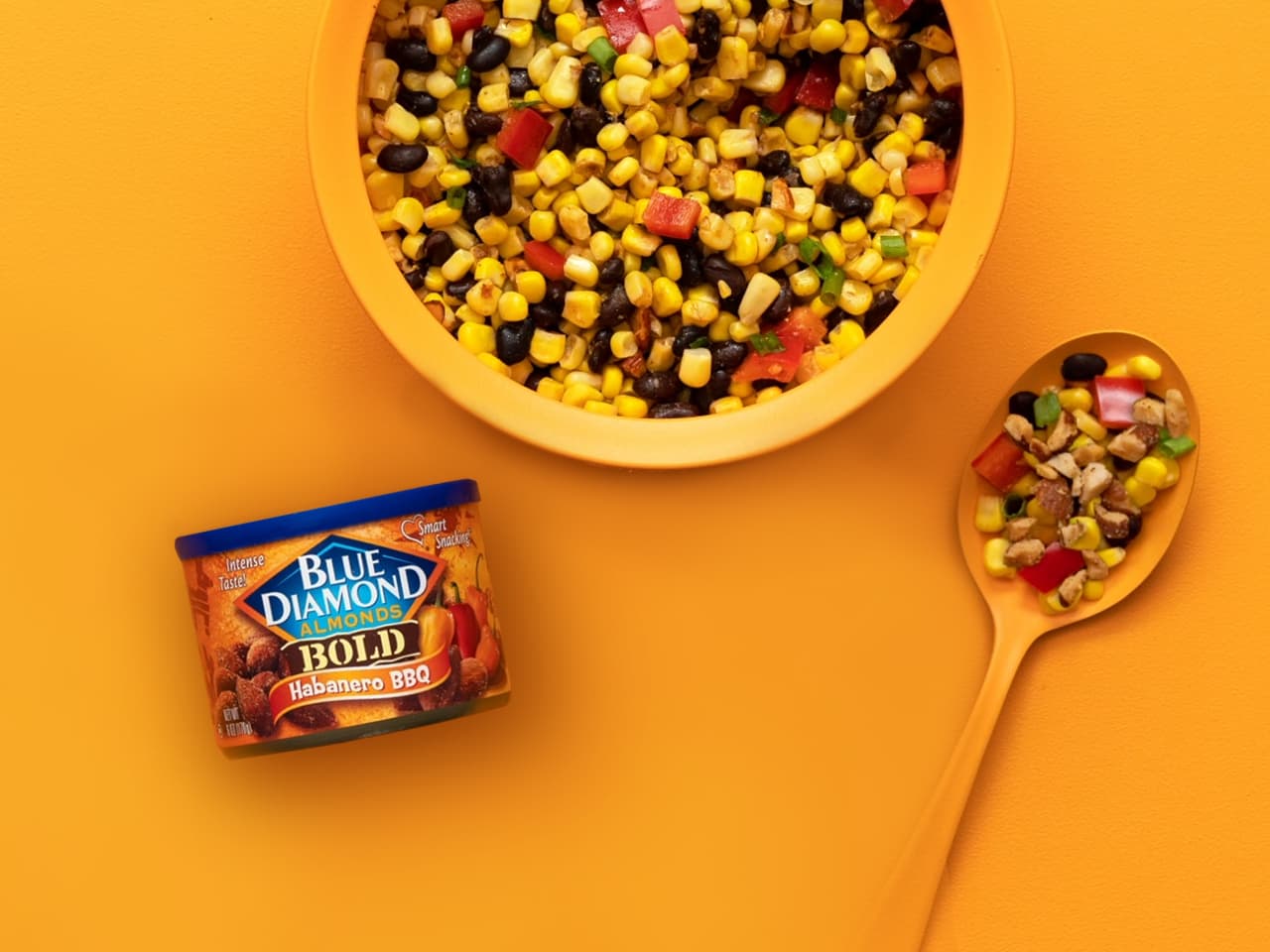A bowl of corn and black bean salad sits on an orange surface beside a spoonful of the salad and a can of Blue Diamond Bold Habanero BBQ Almonds.