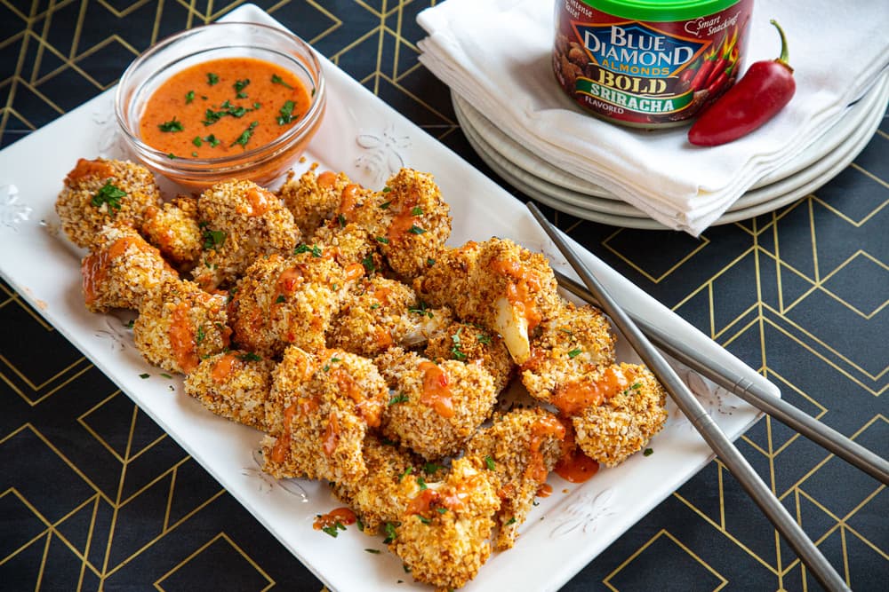 Plate of breaded cauliflower bites drizzled with orange sauce, served with dip, chopsticks, and a can of Blue Diamond Sriracha almonds on stacked white plates.