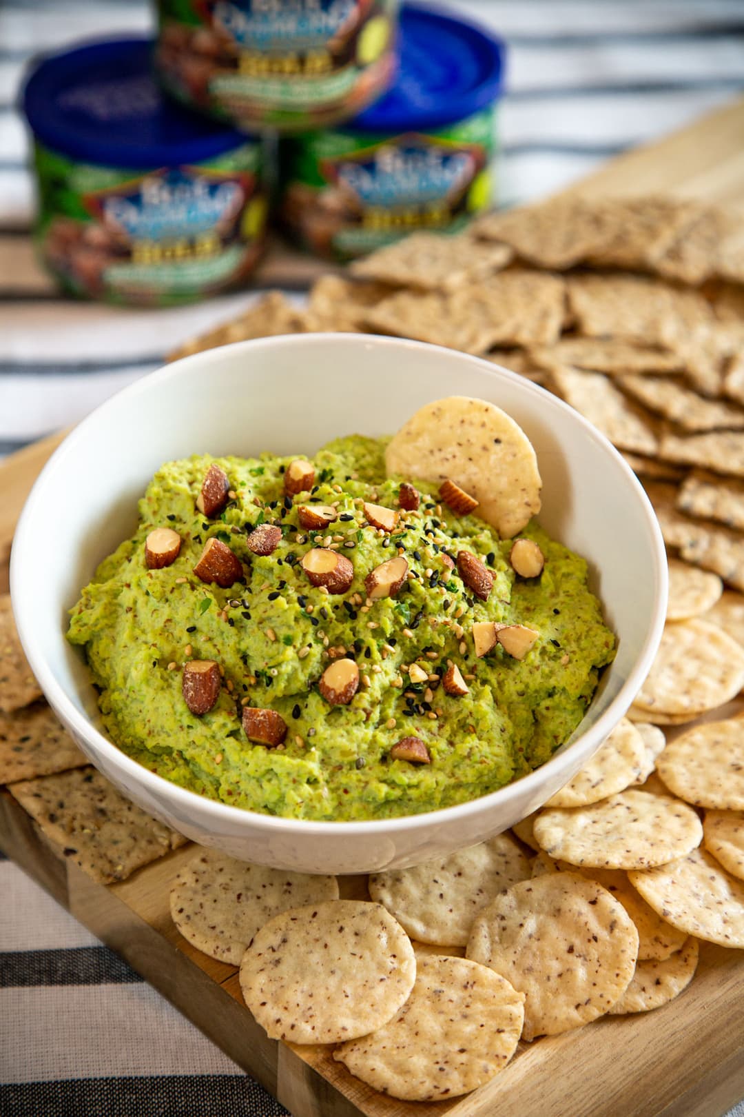 A white bowl of green dip topped with chopped nuts is surrounded by round crackers on a wooden board, with more crackers and snack containers in the background.
