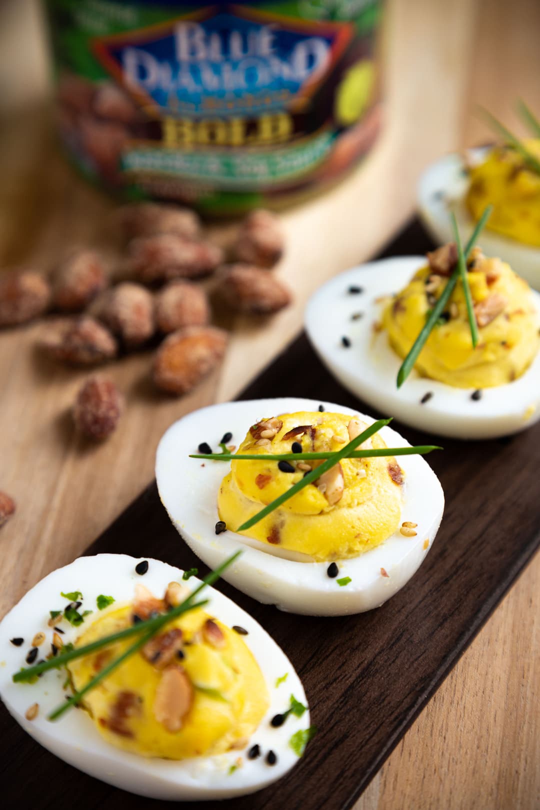 Deviled eggs garnished with herbs, seeds, and sliced almonds are arranged on a dark tray, with whole almonds and an almond container in the background.
