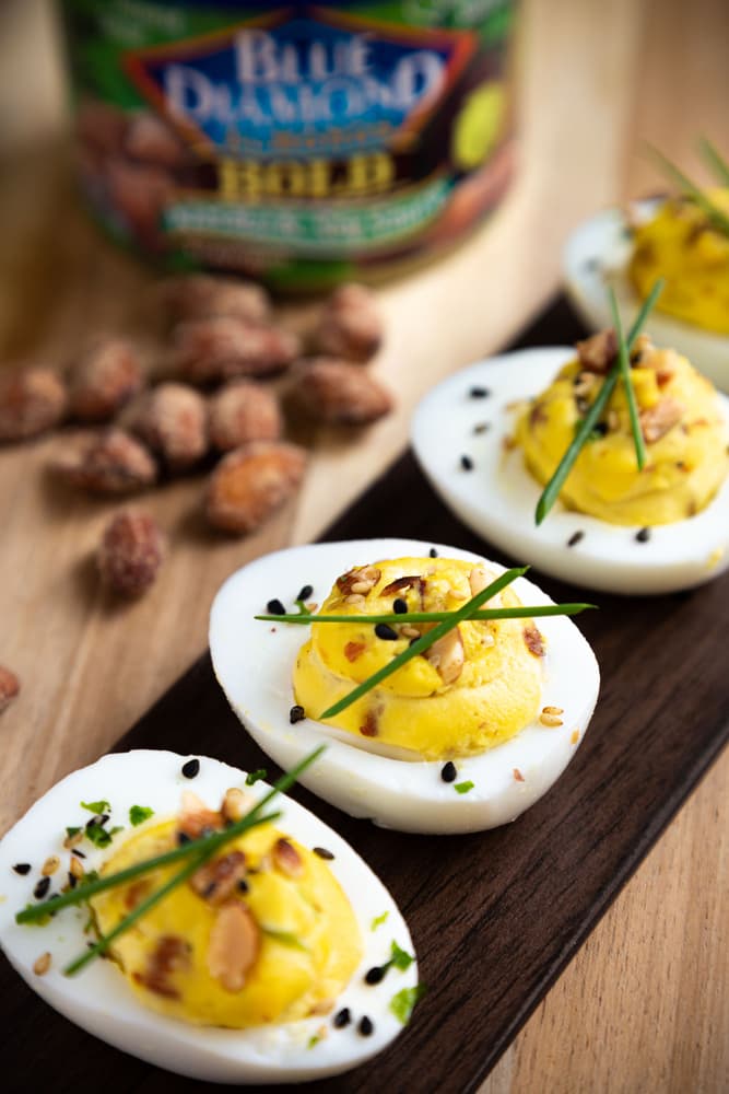 Deviled eggs garnished with herbs, seeds, and sliced almonds are arranged on a dark tray, with whole almonds and an almond container in the background.