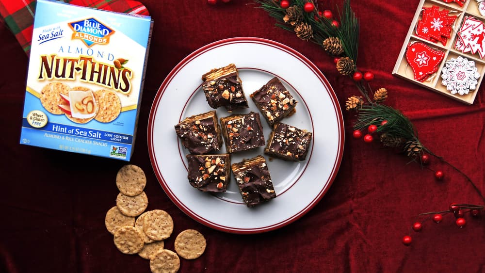 A plate with nine chocolate almond bars sits on a red tablecloth, surrounded by Nut-Thins crackers, a Nut-Thins box, pine branches, pinecones, and decorated holiday cookies.