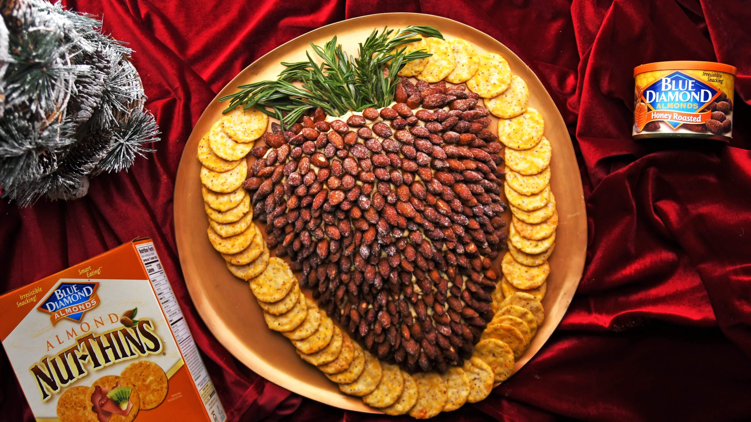 A cheese ball shaped like a pinecone, covered with almonds, is garnished with rosemary and surrounded by crackers on a gold platter atop a red cloth. Nut-Thins and almonds are nearby.