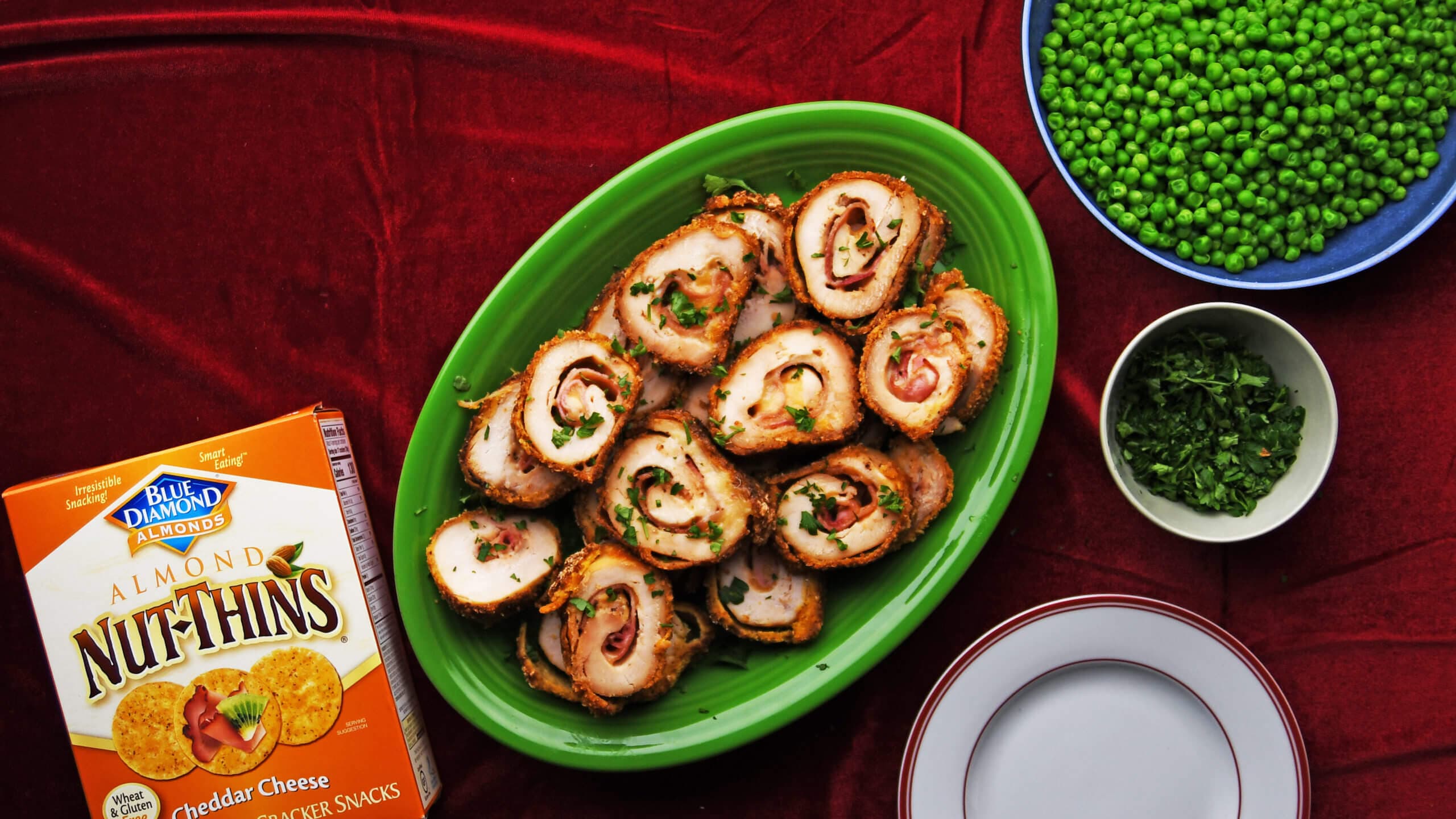 A green platter with sliced stuffed chicken rolls, a bowl of green peas, a bowl of chopped herbs, plates, and a box of Almond Nut-Thins on a red tablecloth.