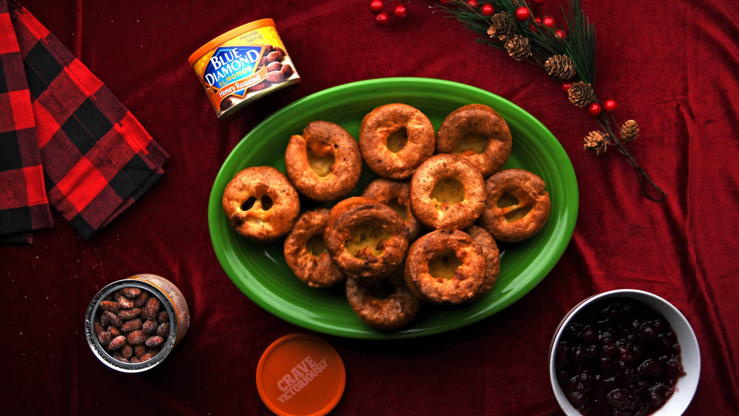 A green plate with Yorkshire puddings on a red tablecloth, surrounded by a can of Blue Diamond almonds, seasoned nuts, cranberries, and holiday decorations.