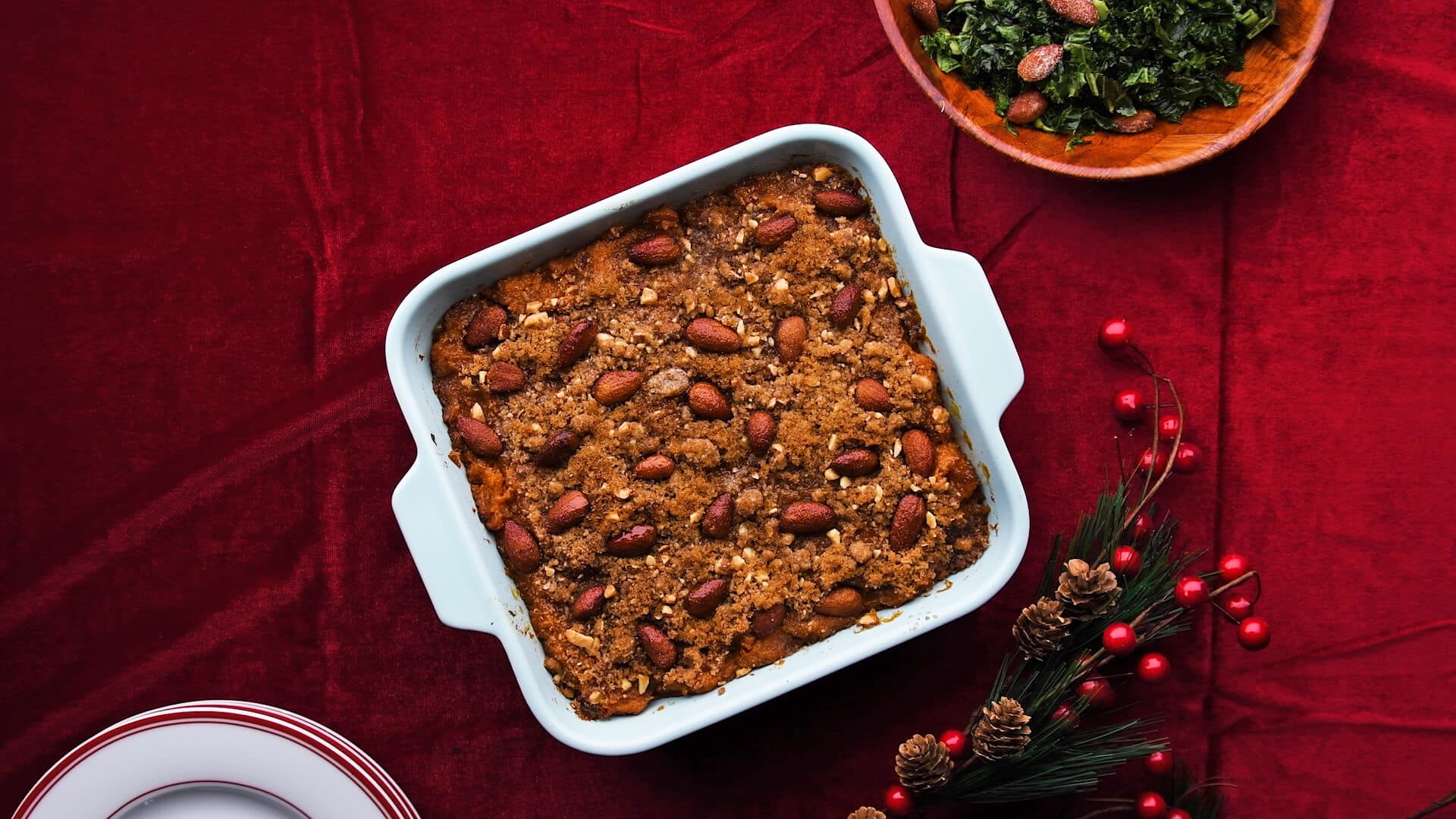 A square baked dessert topped with whole almonds sits in a white dish on a red tablecloth, next to a plate of greens and holiday-themed decorations.