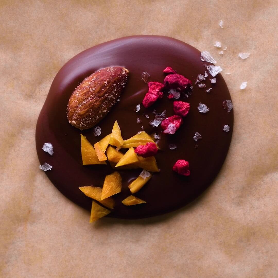 A round pool of chocolate topped with an almond, freeze-dried raspberries, chopped dried fruit, and sea salt flakes on parchment paper.
