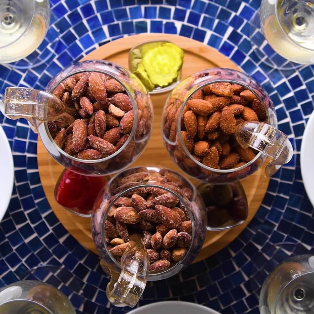 Three glass jars of seasoned almonds on a wooden tray, surrounded by plates and glasses, with pickles and a red condiment on a blue mosaic table.